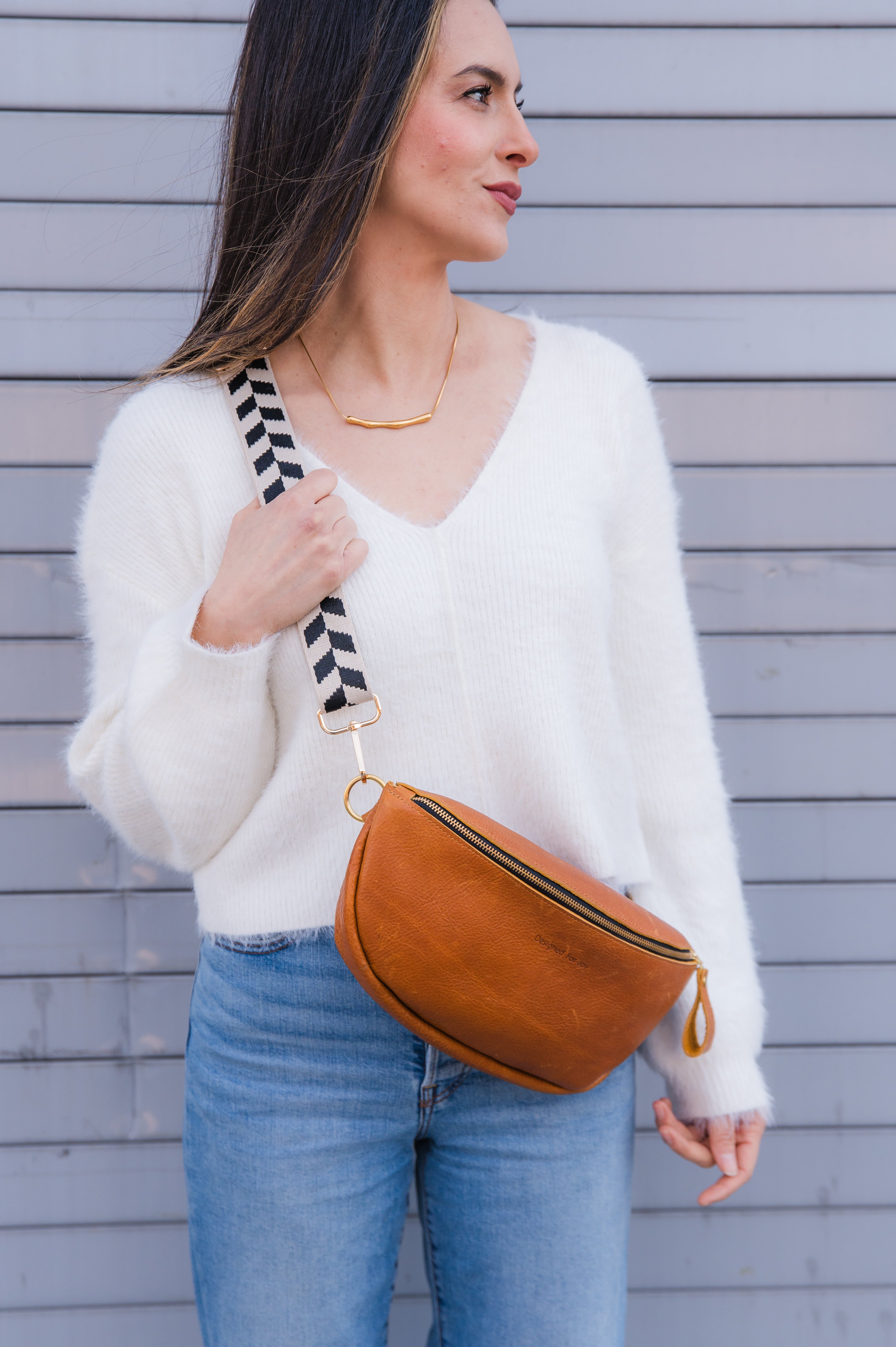 Person wearing a caramel brown crossbody sling handbag while holding the strap in one hand and looking to the side