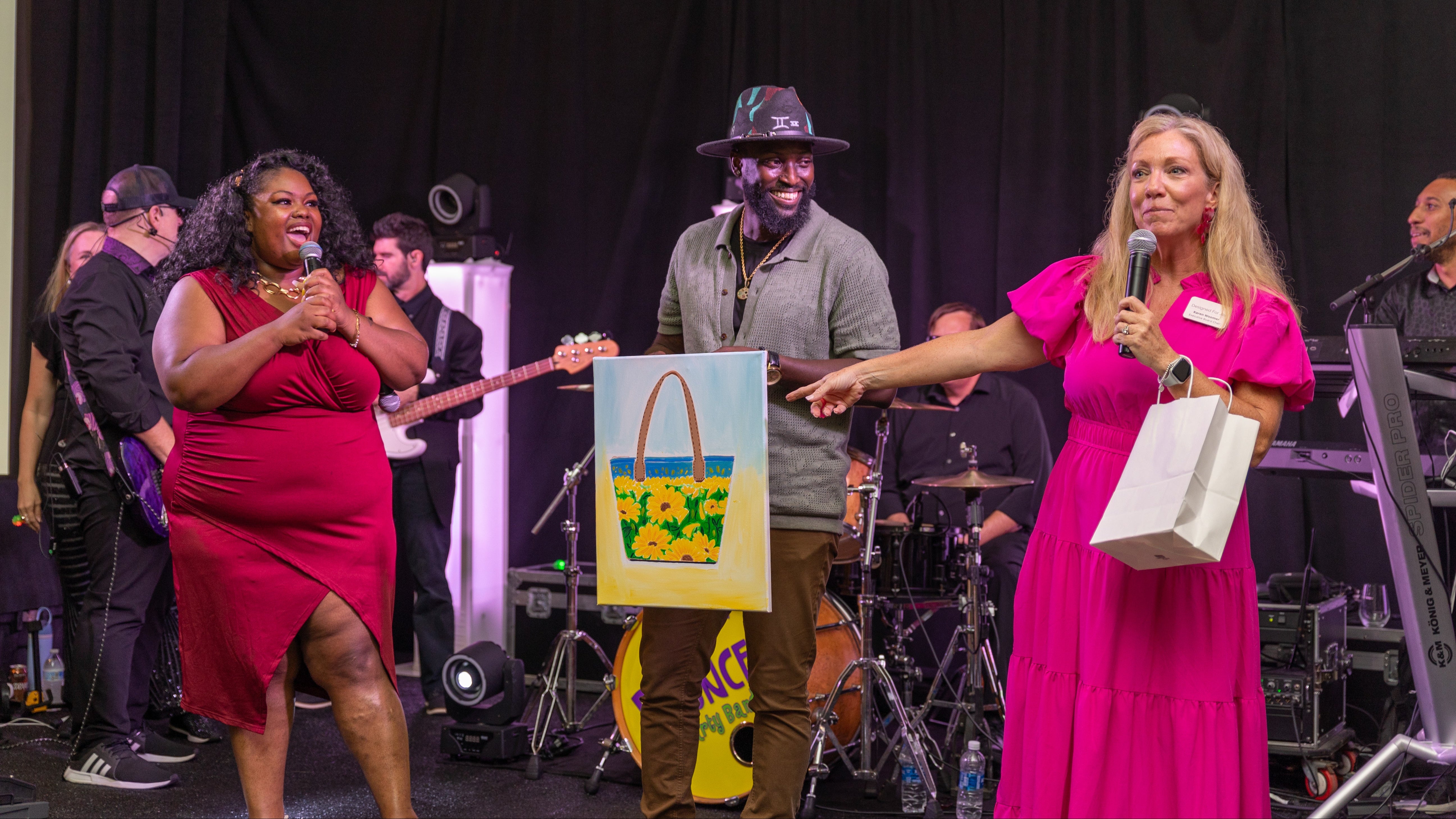 Two women on stage with a band, one holding a microphone and a colorful bag, the other holding a gift box.