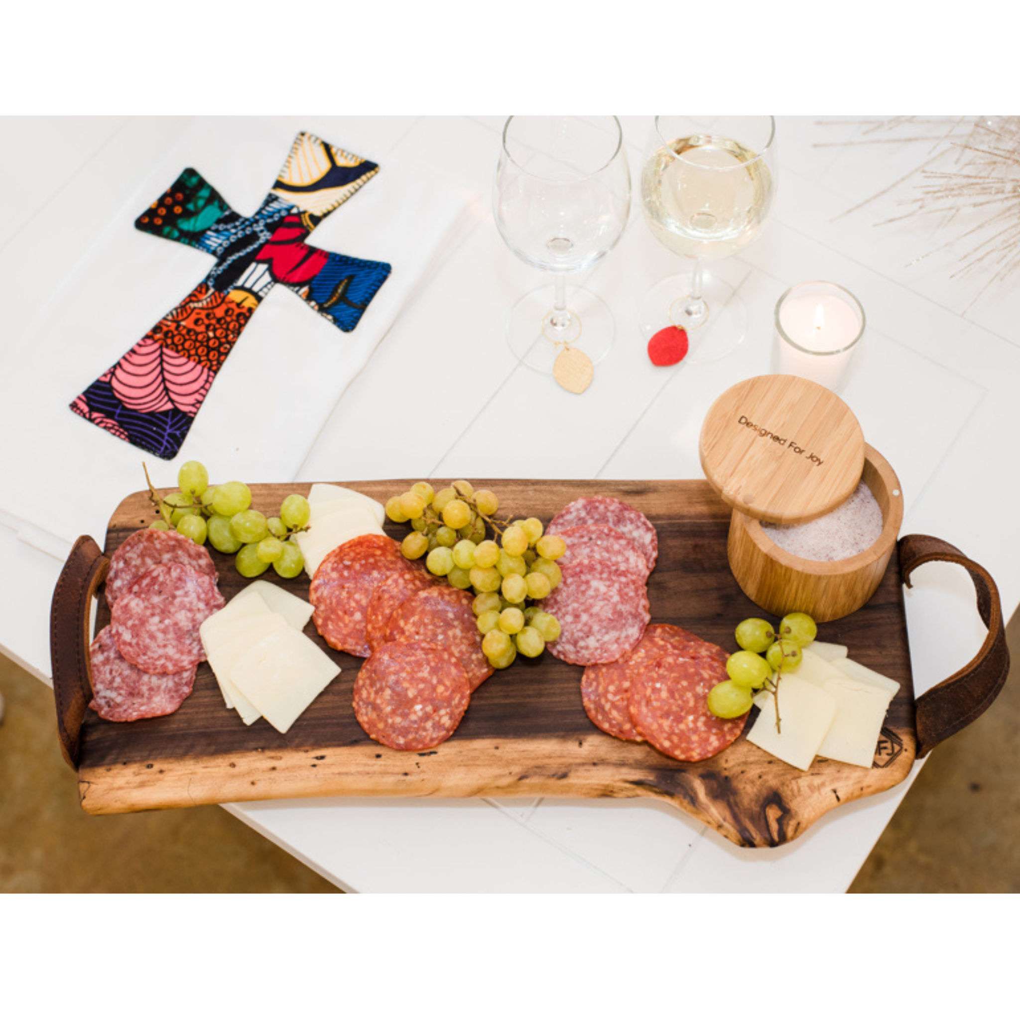 A black walnut wood charcuterie board on a table with a spread of cheese, grapes, and meats, next to wine glasses and a fabric towel