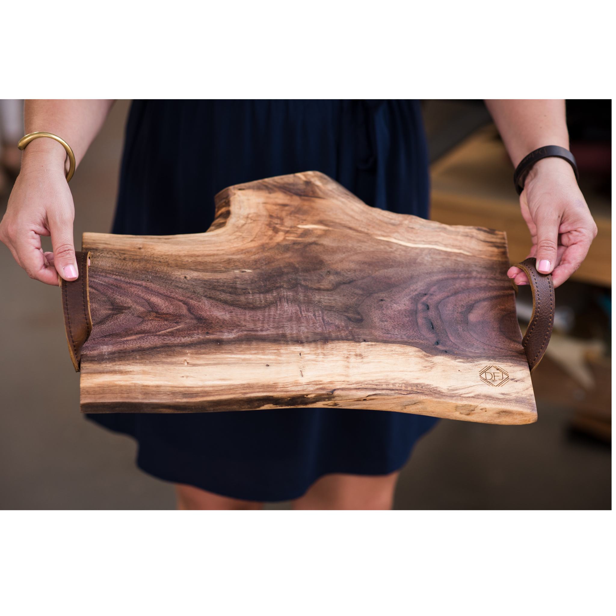 A person holding a live-edge charcuterie board made of black walnut wood with leather handles