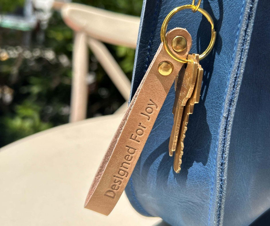 Brown keychain with keys and leather tag with 'Designed for Joy' text on a blue bag in an outdoor setting