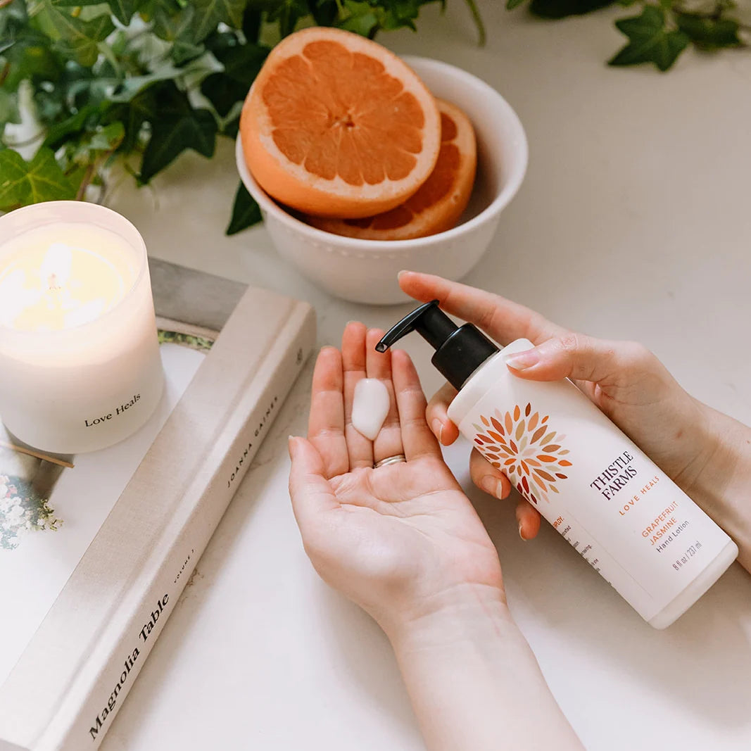 A hand holding a pump lotion labeled 'Grapefruit Jasmine' while the other has lotion on it with candles, a book, fruit, and leaves in the background