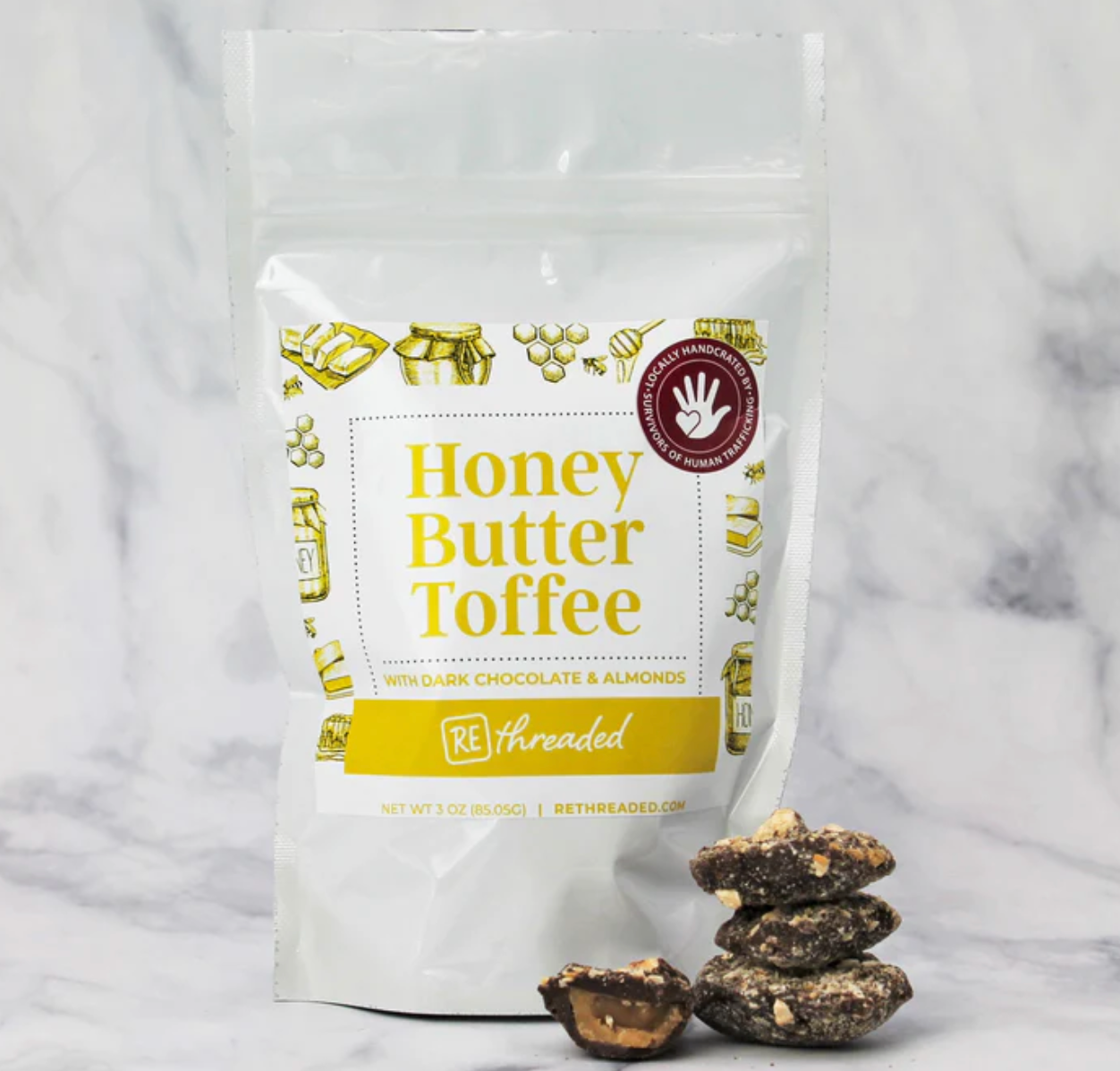 A white bag labeled 'Honey Butter Toffee with Dark Chocolate & Almonds', displayed alongside pieces of toffee on a marble background
