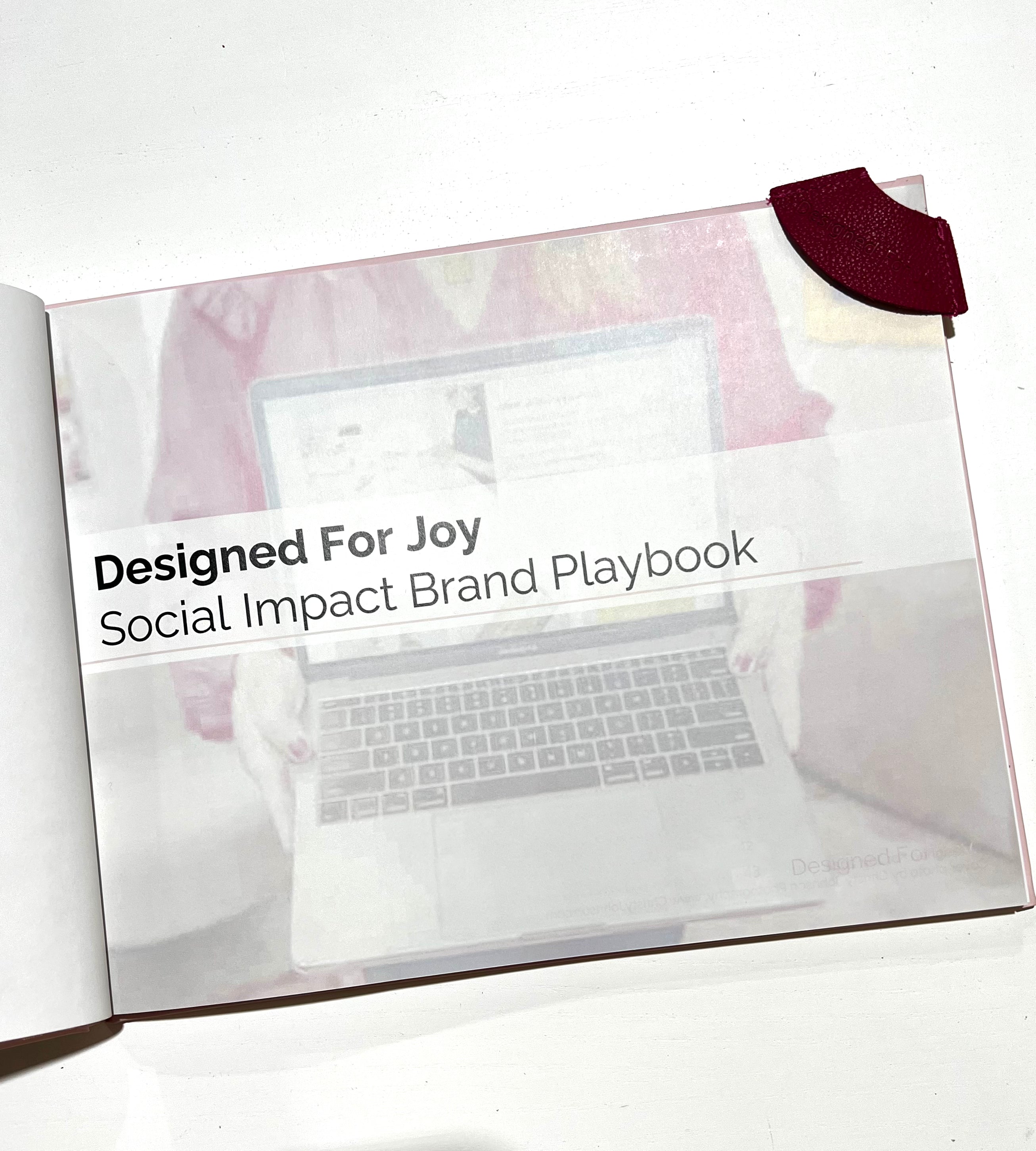An open book showing a page with the text 'Designed for Joy Social Impact Brand Playbook' with a pink leather bookmark on the corner