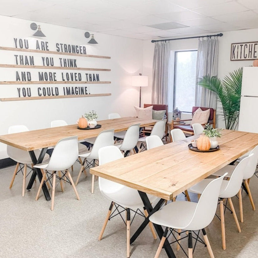 Well-lit room with two large wooden tables accompanied by white chairs and decorative items such as a framed quote on the wall that reads 'YOU ARE STRONGER THAN YOU THINK AND MORE LOVED THAN YOU COULD IMAGINE.'