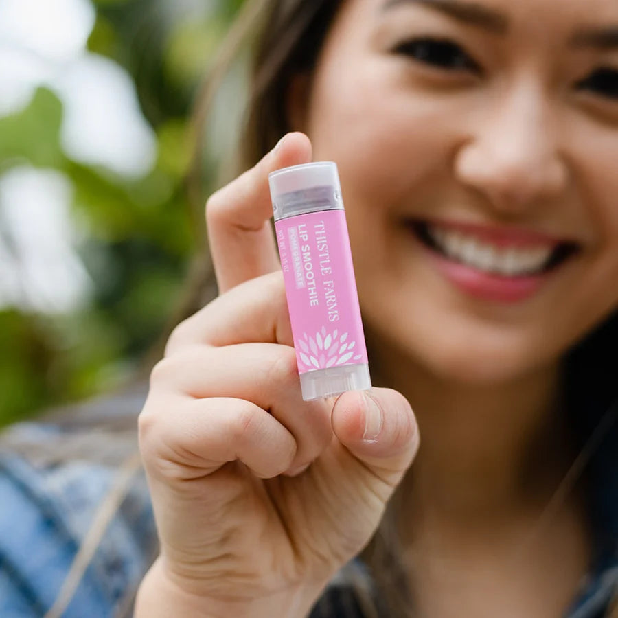 A person holding a stick of 'Thistle Farms Lip Smoothie Pomegranate' against a blurred background