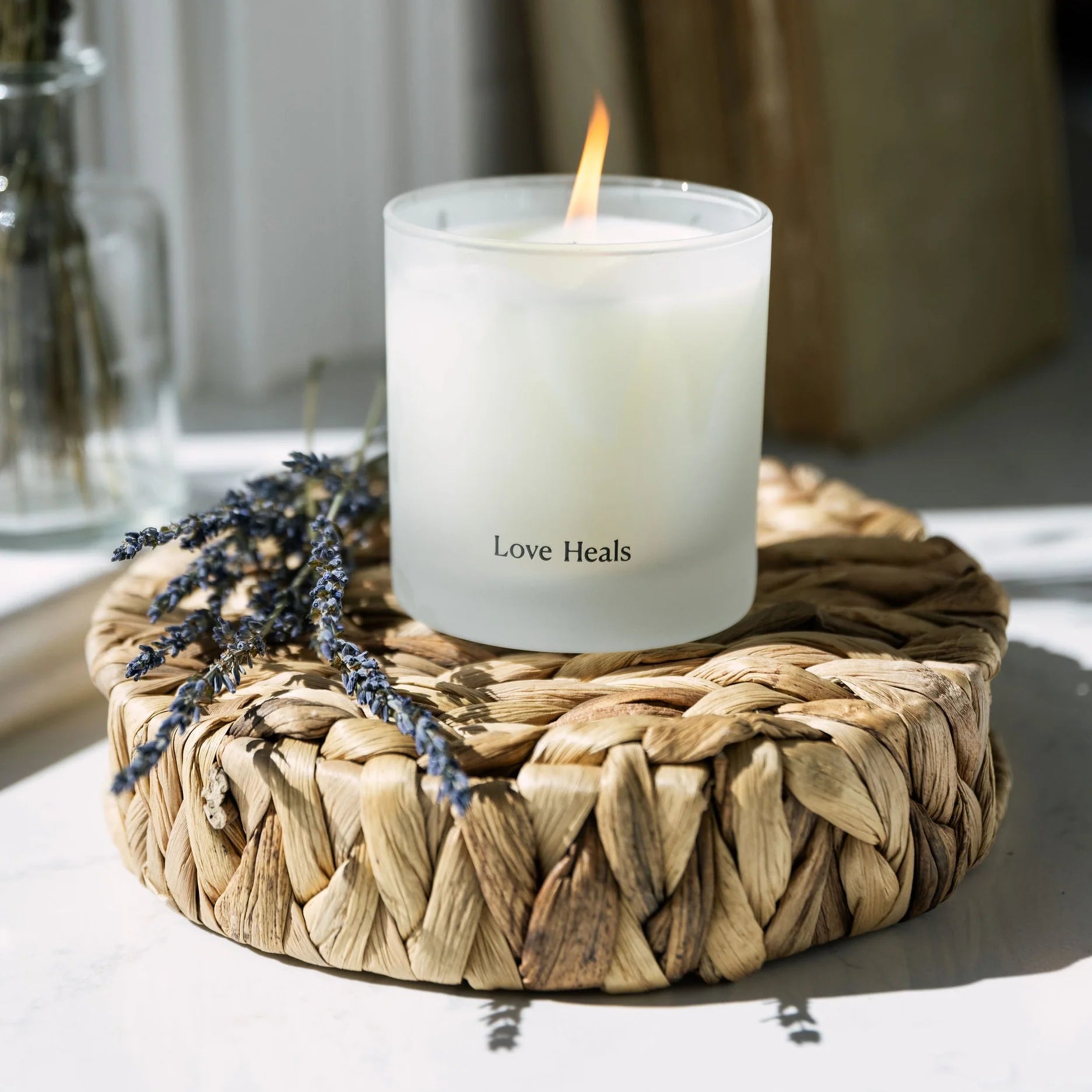A Thistle Farms soy candle, branded with 'Love Heals', showcased on a round wooden display next to dried lavender