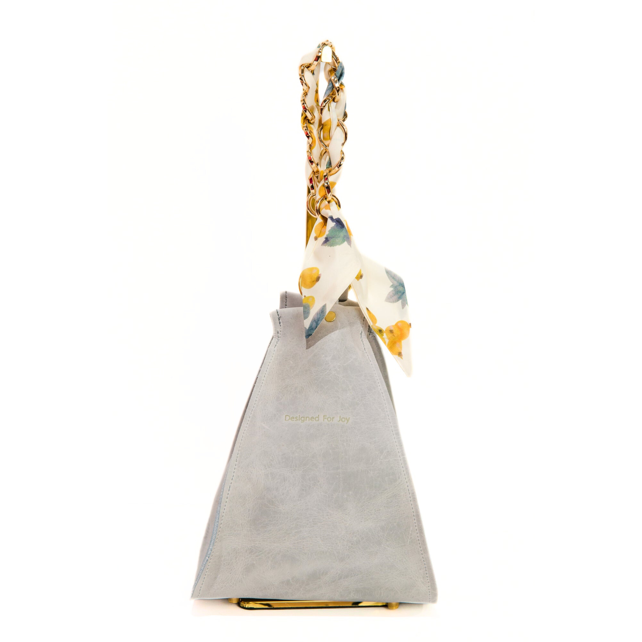 Gray bucket handbag with a gold chain wrist strap that has a removable scarf and 'Designed for Joy' text, displayed on a white background