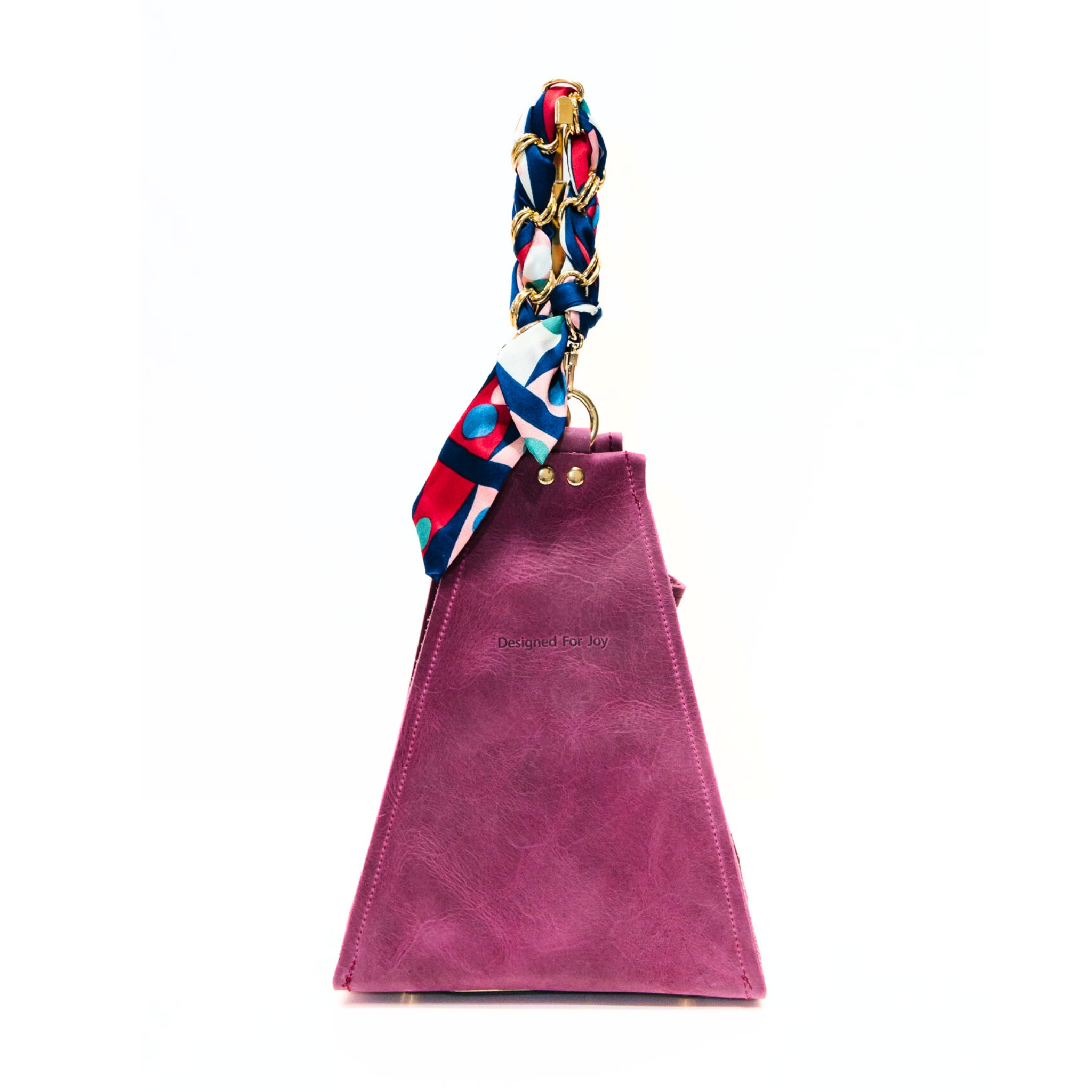 Raspberry pink bucket handbag with a gold chain wrist strap that has a removable scarf and 'Designed for Joy' text, displayed on a white background