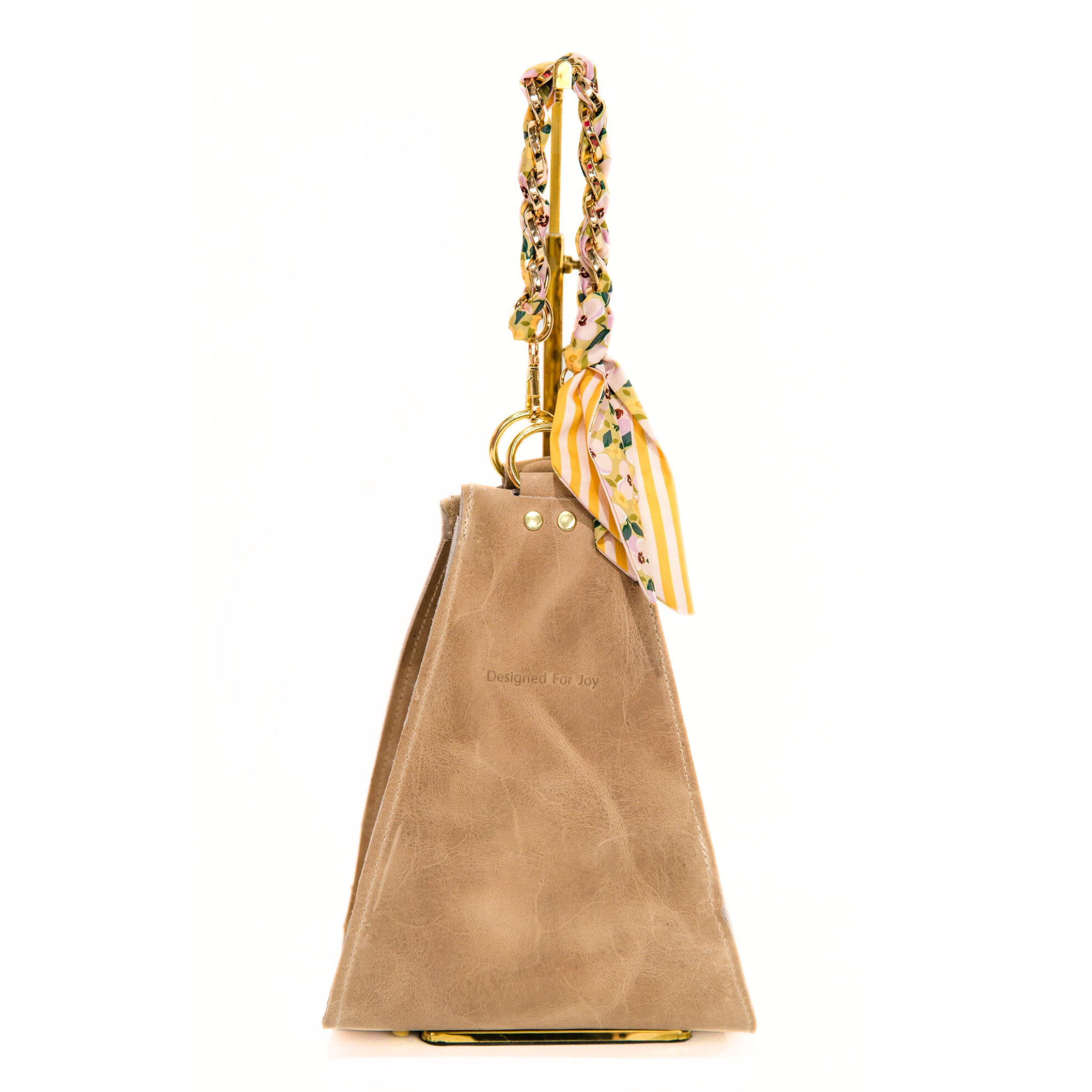 Tan bucket handbag with a gold chain wrist strap that has a removable scarf and 'Designed for Joy' text, displayed on a white background