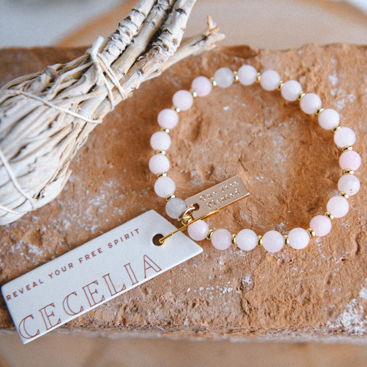 Rose quartz beaded bracelet with gold colored spacer beads, and tags with 'One Tree Planted' and 'Reveal Your Free Spirit Cecelia' text, displayed on a stone surface