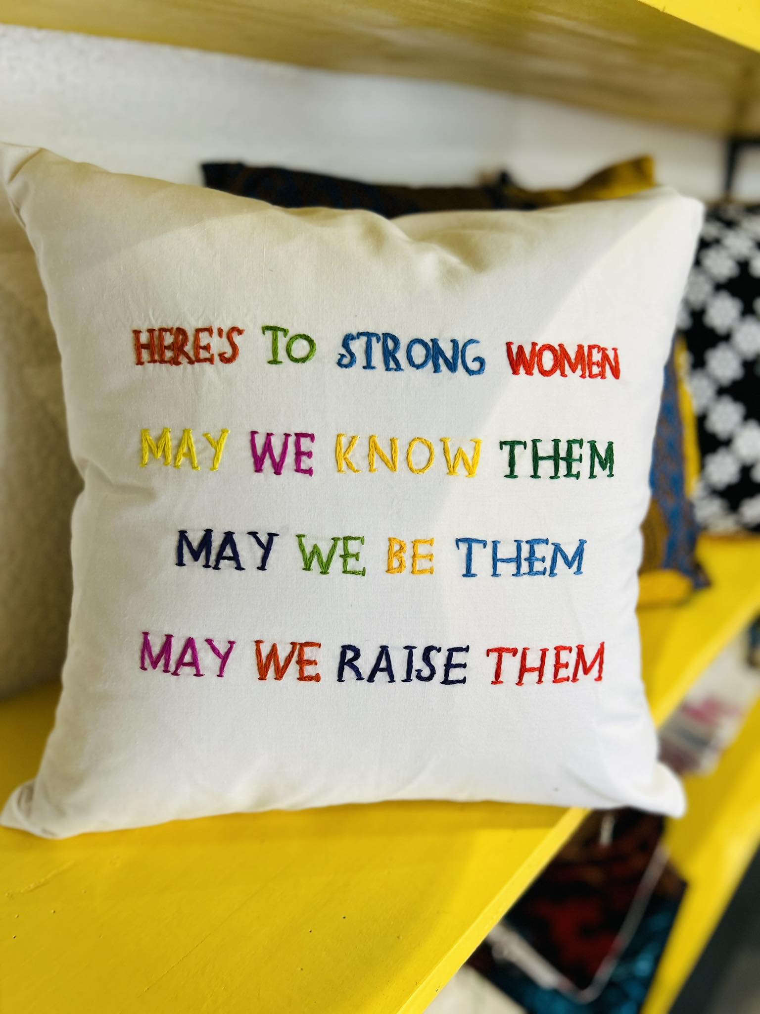 Decorative white pillow with the message 'HERE'S TO STRONG WOMEN MAY WE KNOW THEM MAY WE BE THEM MAY WE RAISE THEM' printed in multicolored letters