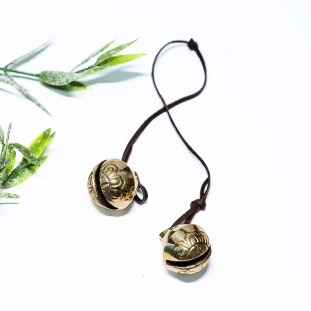 Two gold-colored jingle bells tied together with soft deerskin leather, displayed on a white background with green leaves to the side