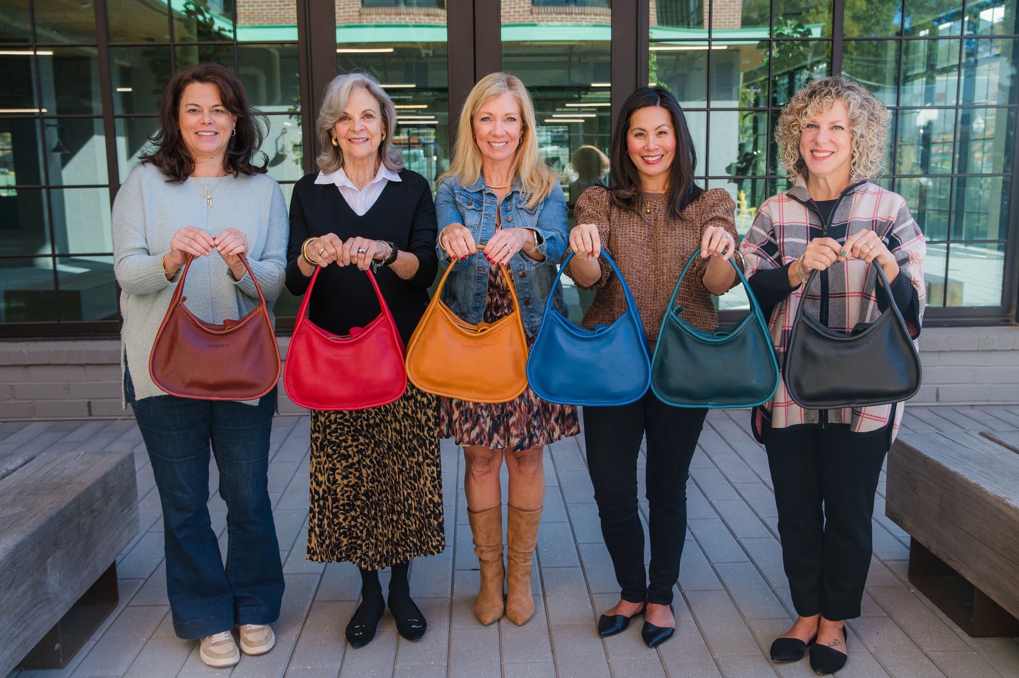 Line of women holding various shoulder handbags in the colors cinnamon brown, pink, caramel brown, blue, teal, and black, in an outdoor setting
