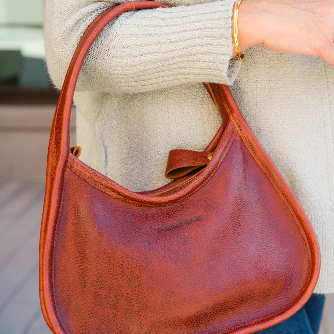 Close-up of a person holding a cinnamon brown shoulder handbag in an outdoor setting