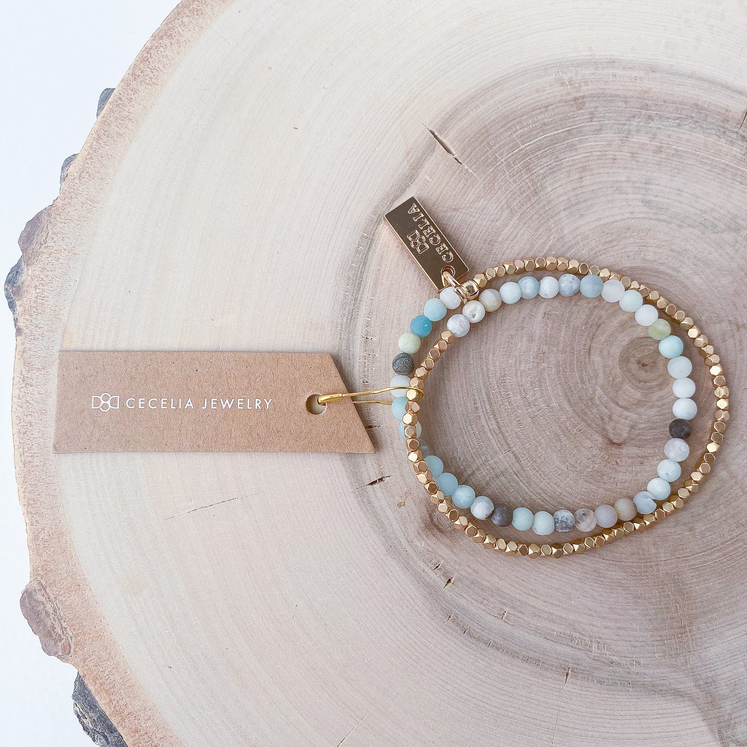 A half amazonite, half matte gold-colored beaded bracelet on a wooden surface with a tag that has 'Cecelia Jewelry' text