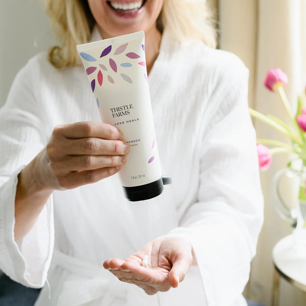 Person holding a tube of Thistle Farms Hand and Body Lotion labeled 'Love Heals', with flowers in the background