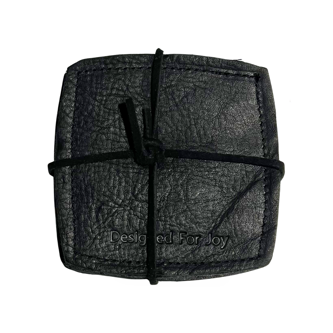 Black leather coasters with 'Designed for Joy' imprinted on them, held together with leather lacing, on a white background