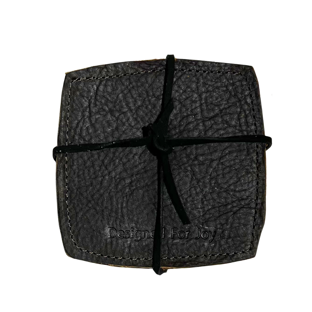 Brown leather coasters with 'Designed for Joy' imprinted on them, held together with leather lacing, on a white background