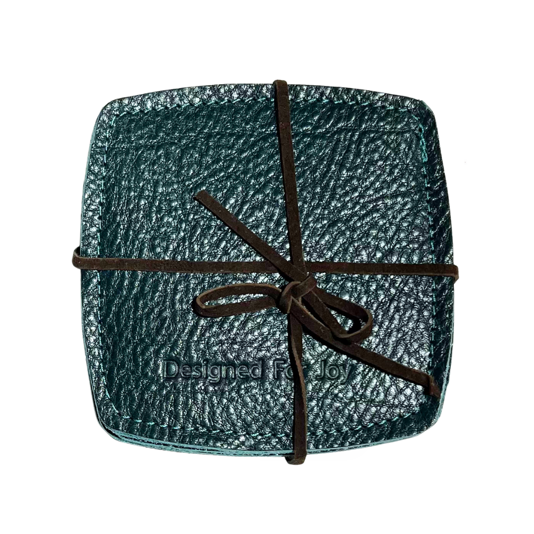 Teal leather coasters with 'Designed for Joy' imprinted on them, held together with leather lacing, on a white background