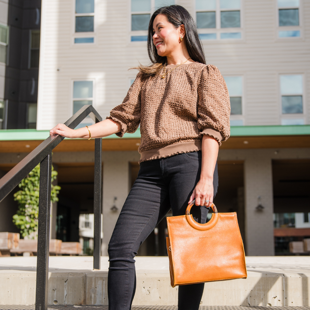 Person holding a caramel brown satchel handbag while standing on stairs in an outdoor setting