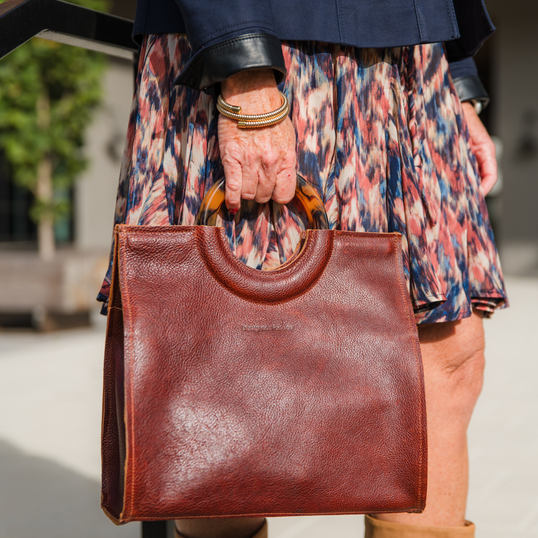 Close-up of a person holding a cinnamon brown satchel handbag in an outdoor setting