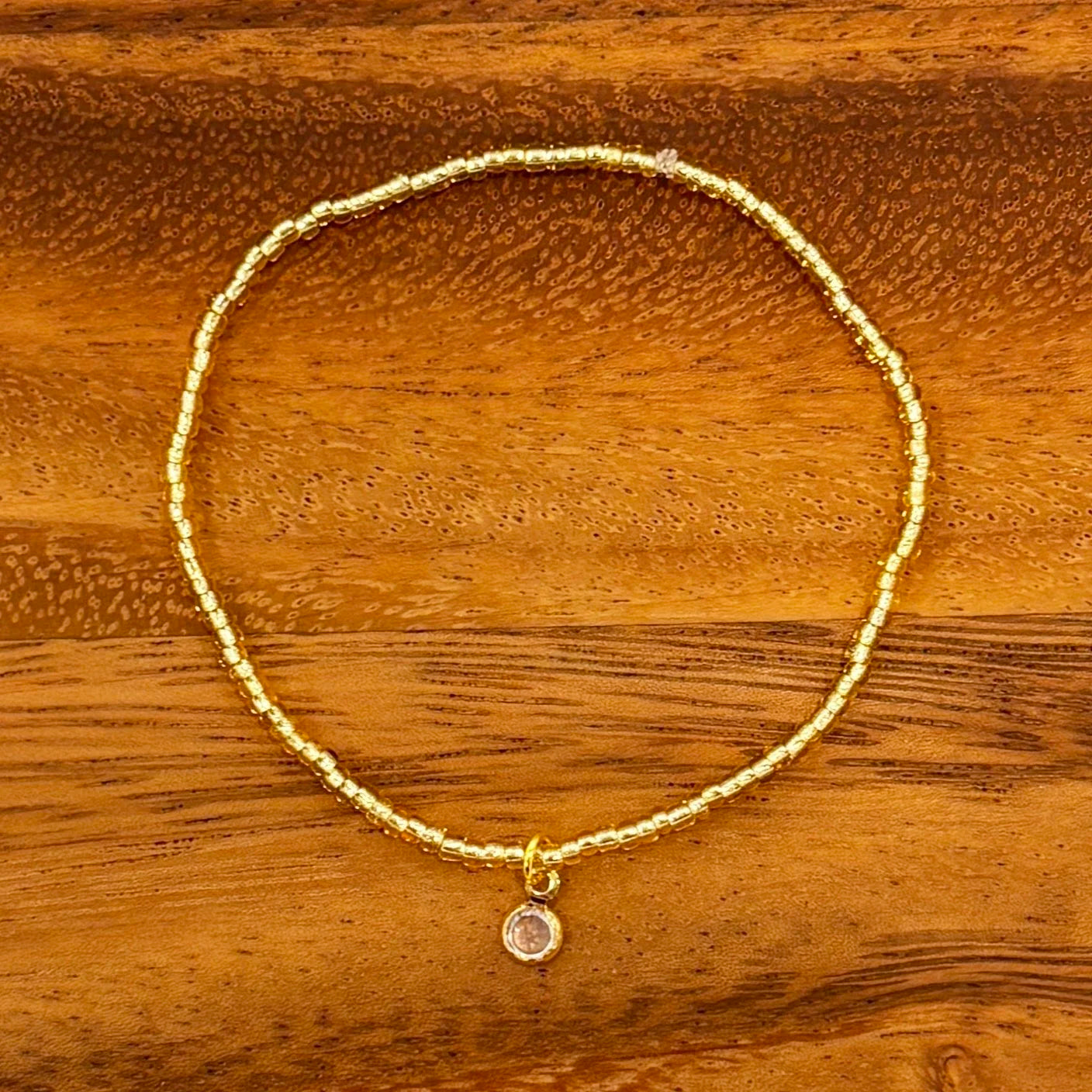 Gold seed beaded bracelet with circular crystal dangle charm, displayed on a wooden background