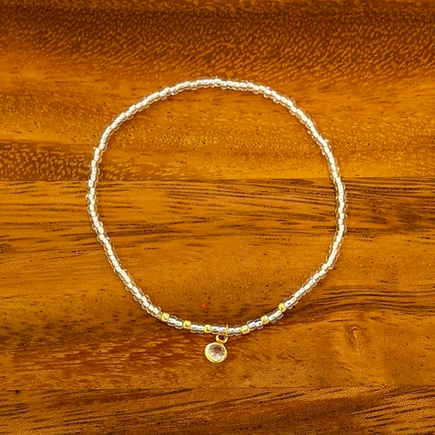 Silver seed beaded bracelet with gold accent beads and circular crystal dangle charm, displayed on a wooden background