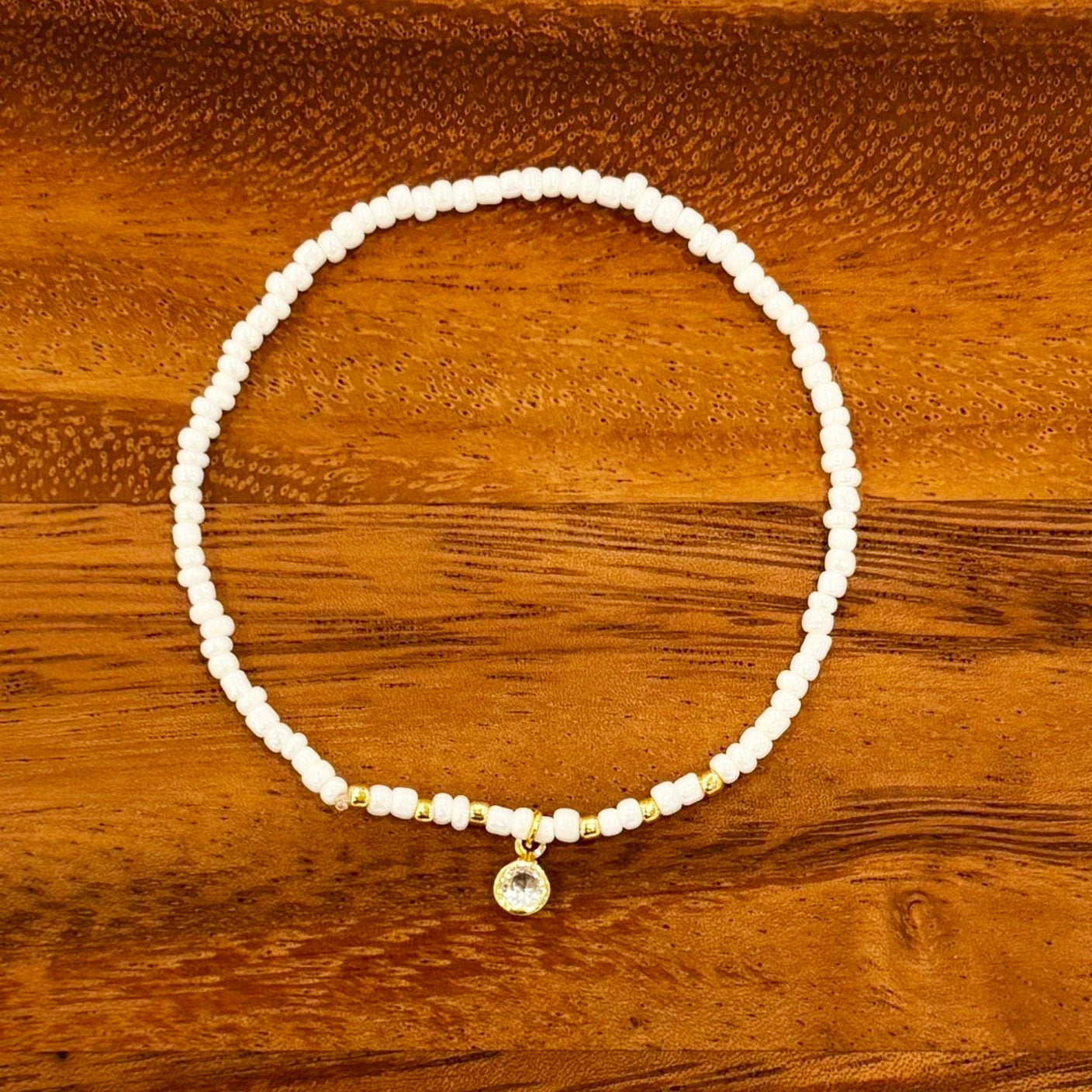 White seed beaded bracelet with gold accent beads and circular crystal dangle charm, displayed on a wooden background