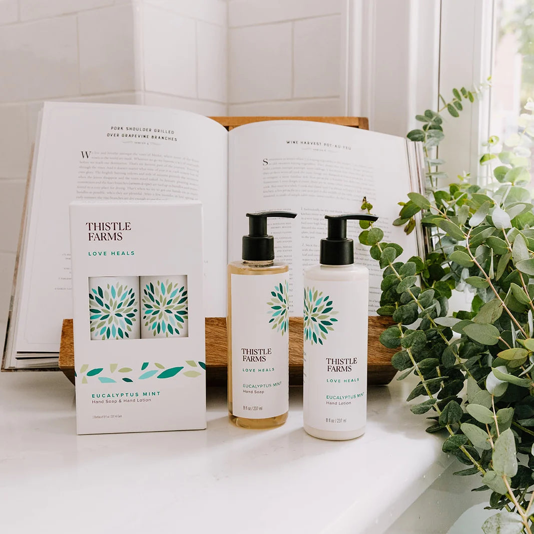 Box of the Thistle Farms Hand Soap and Hand Lotion in eucalyptus mint, next to the individual bottles with a book and a plant in the background