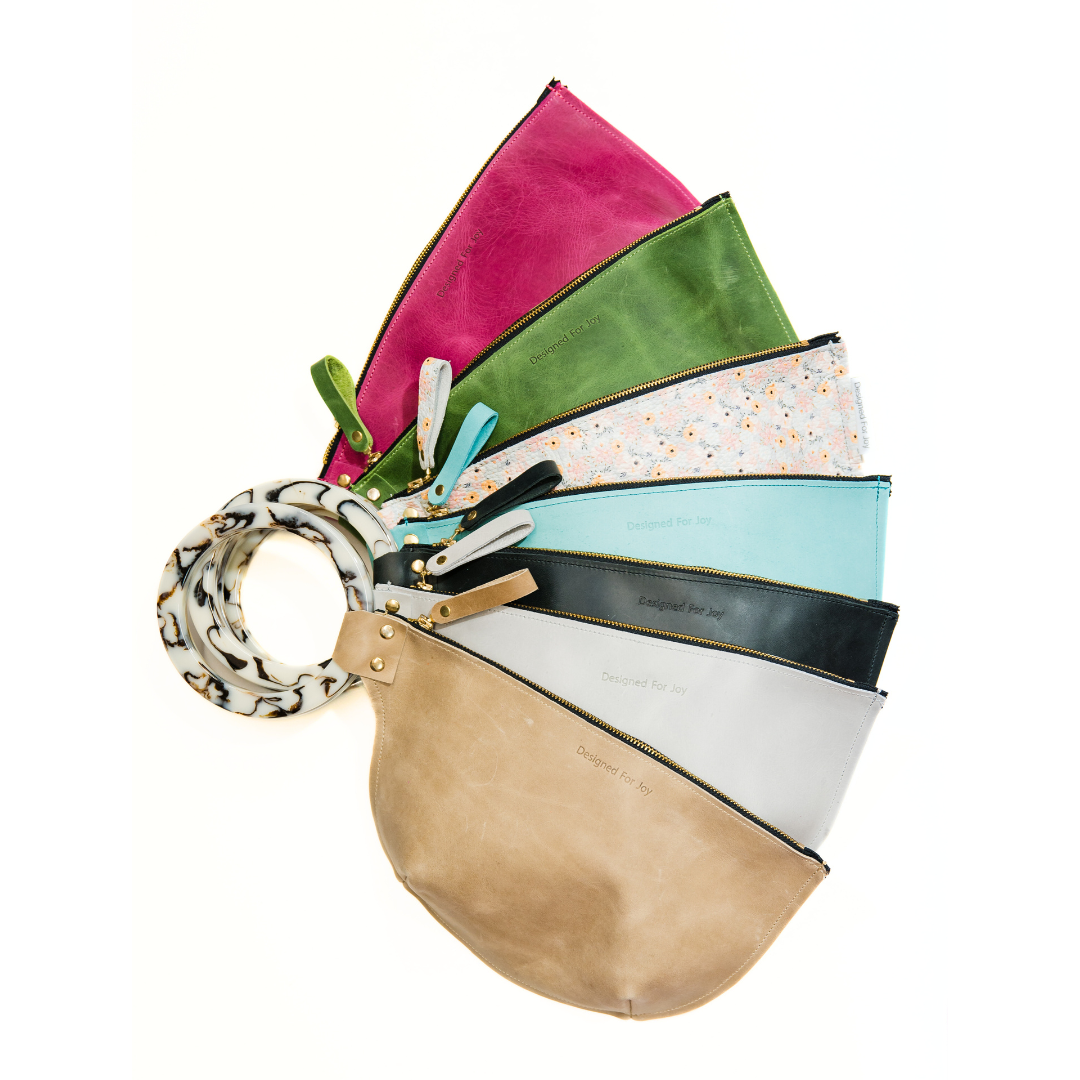 A collection of wristlet handbags in various colors displayed in a fan-like arrangement, featuring a smooth resin handle and a zippered compartment