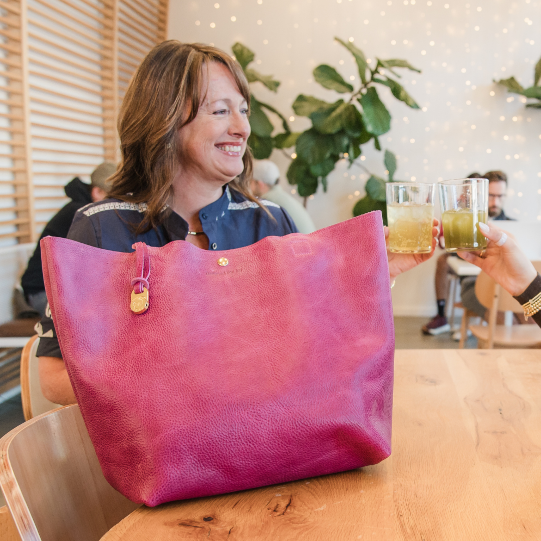 A person clinking glass cups with somebody else in a cafe setting with their large pink tote bag on the table next to them