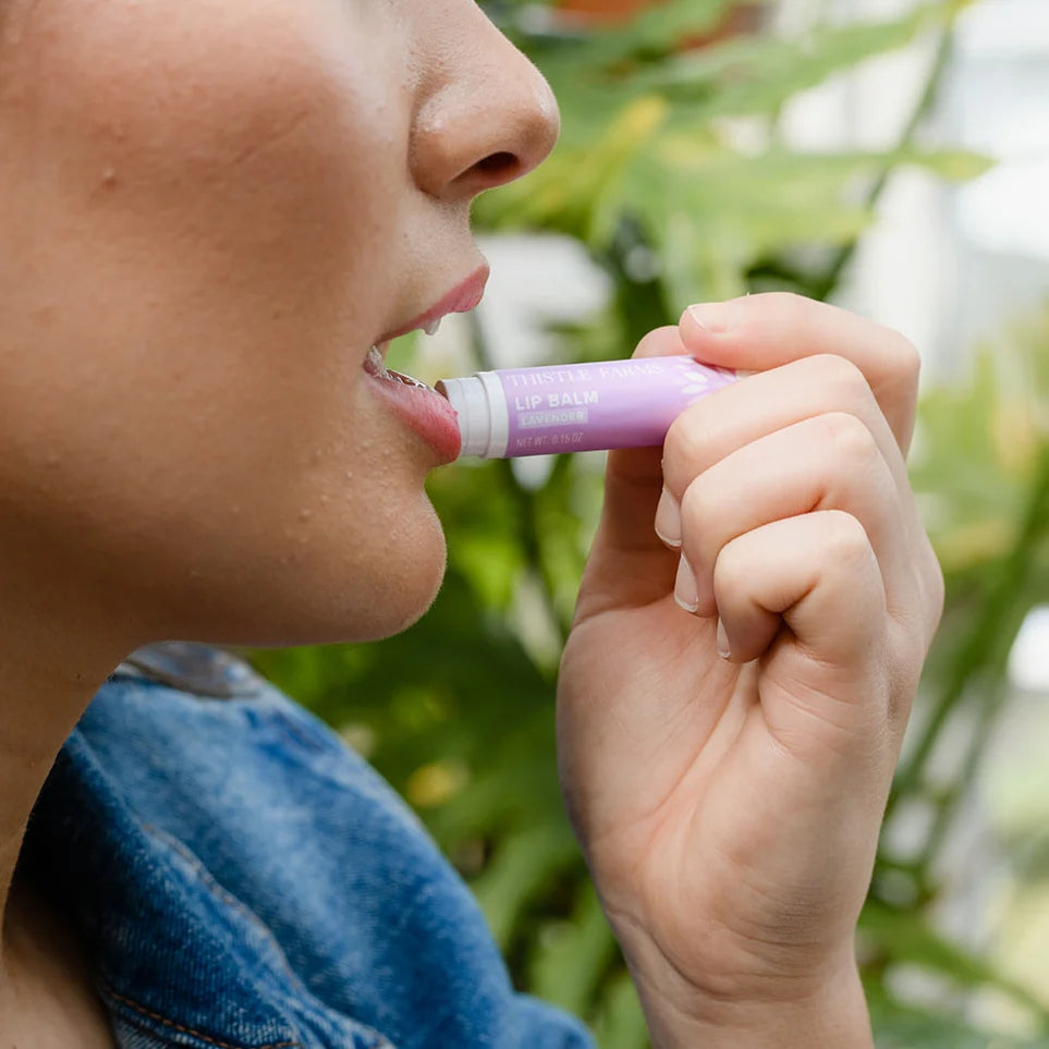Close-up of a person wearing 'Thistle Farms Lip Balm Lavender' in a blurred greenery setting