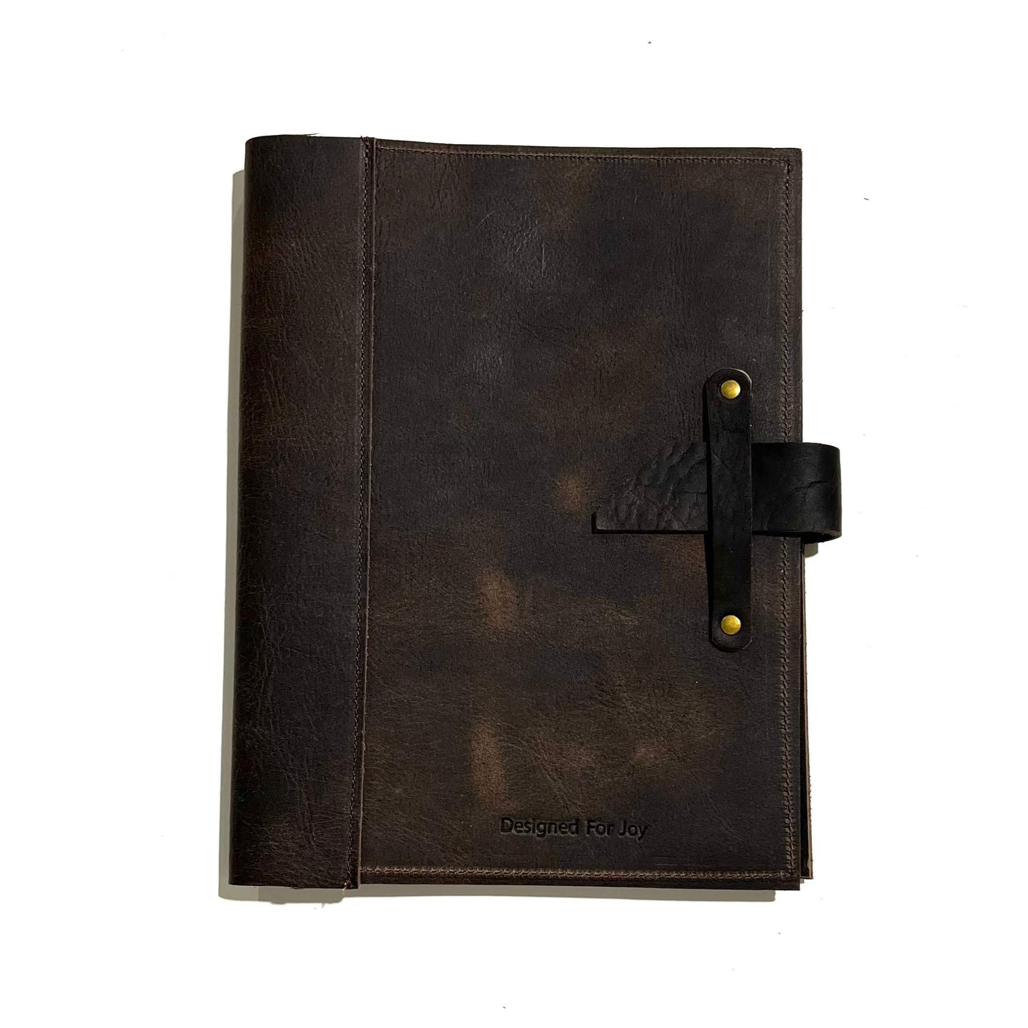 Brown leather journal with a wide leather tab closure and 'Designed for Joy' text, displayed on a white background