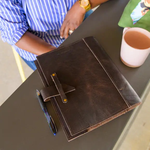 A brown leather journal displayed on a blurred background of a person sitting at a table with coffee and a pen