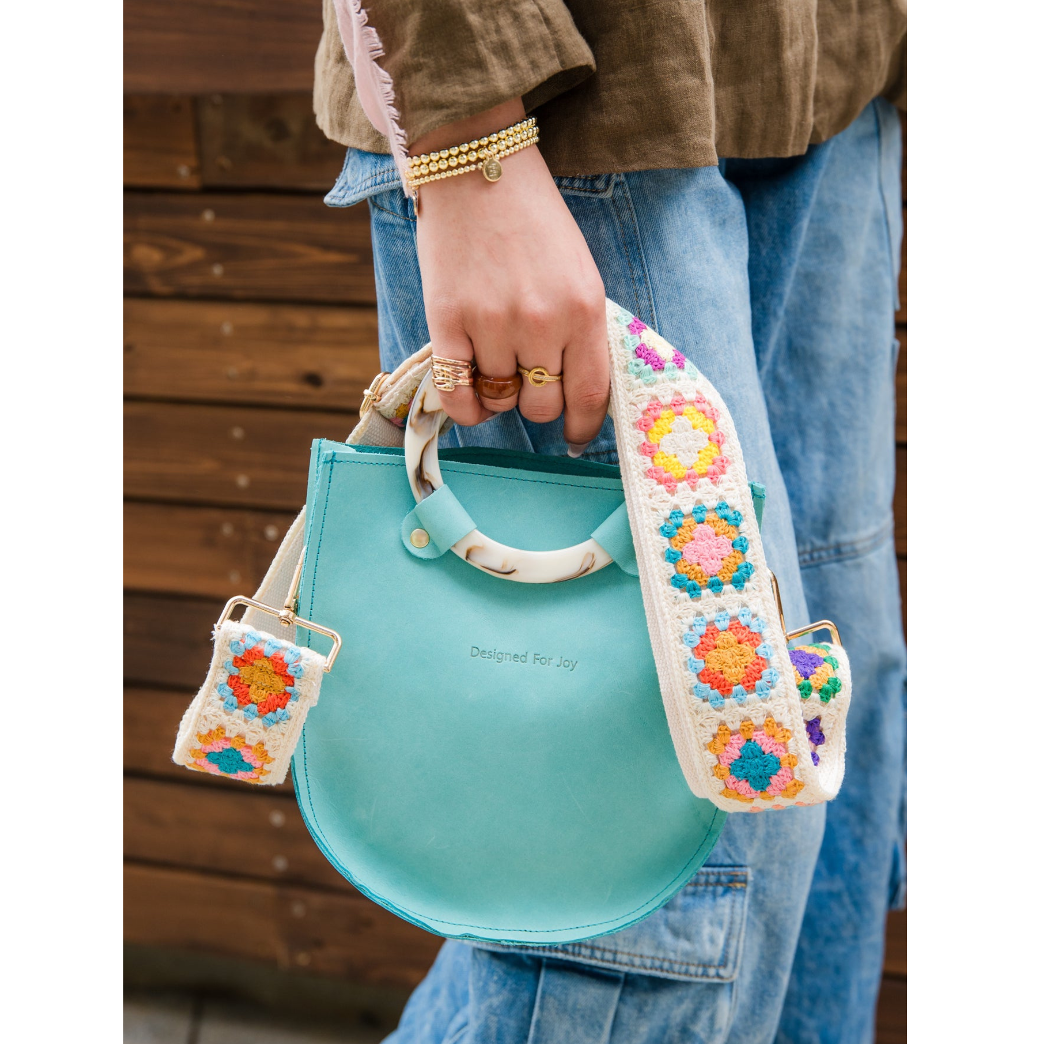 Close-up of a person holding an aqua blue top handle handbag with a colorful crochet crossbody strap attached to it