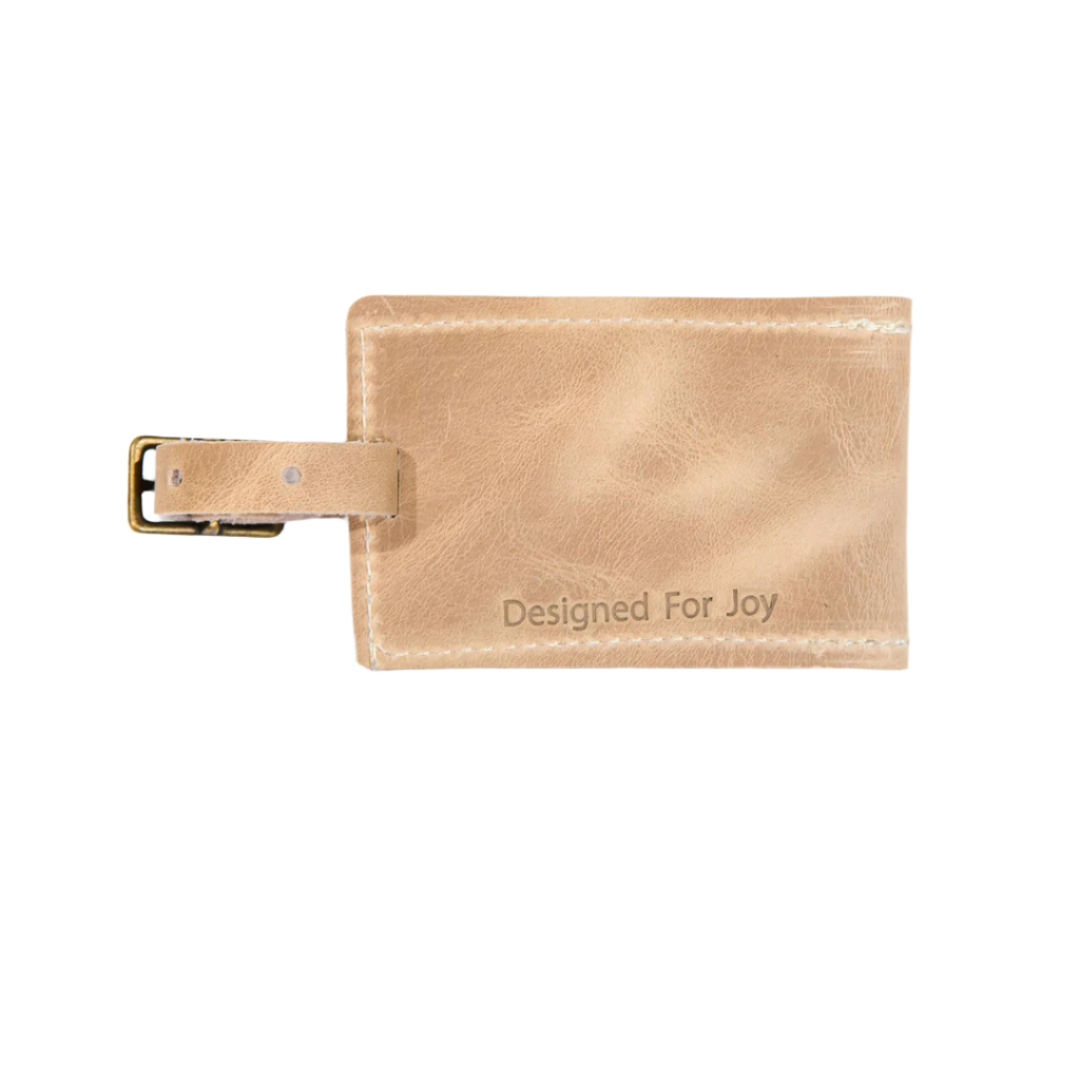 Tan luggage tag with a leather buckled strap and 'Designed for Joy' text, displayed on a white background
