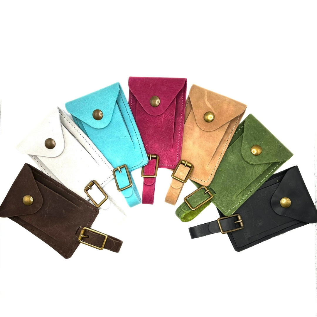 A collection of luggage tags in various colors, including brown, gray, aqua blue, raspberry pink, tan, evergreen, and black, each featuring a leather buckled strap and snap closure
