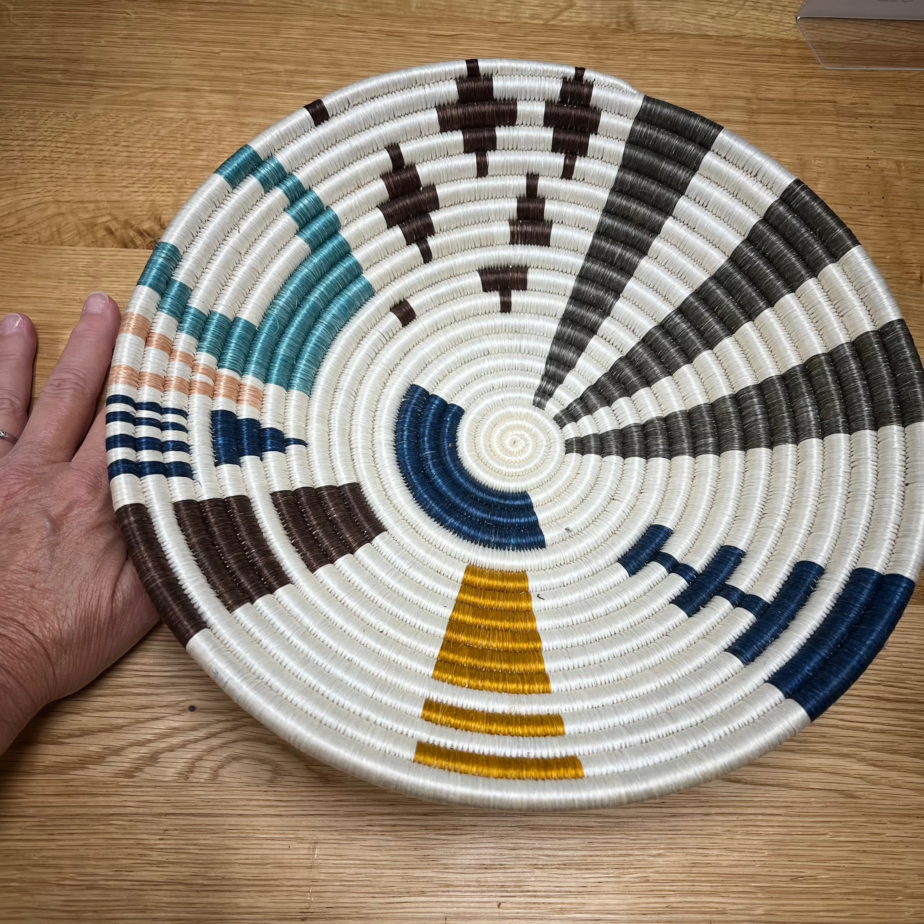 A medium Rwandan basket with a geometric pattern, displayed on a wooden surface next to a hand