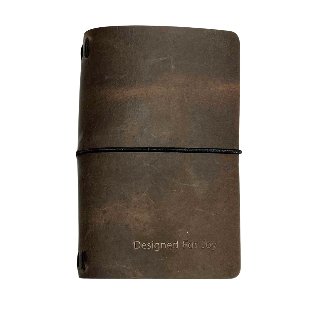 Brown notebook with 'Designed for Joy' text on the cover and a black strap, on a white background