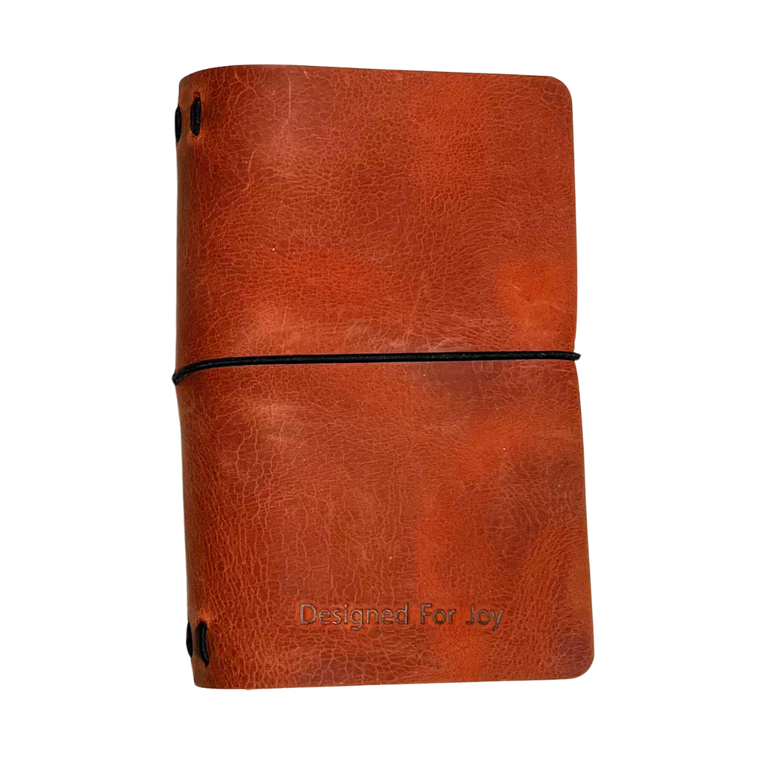 Dark orange notebook with 'Designed for Joy' text on the cover and a black strap, on a white background