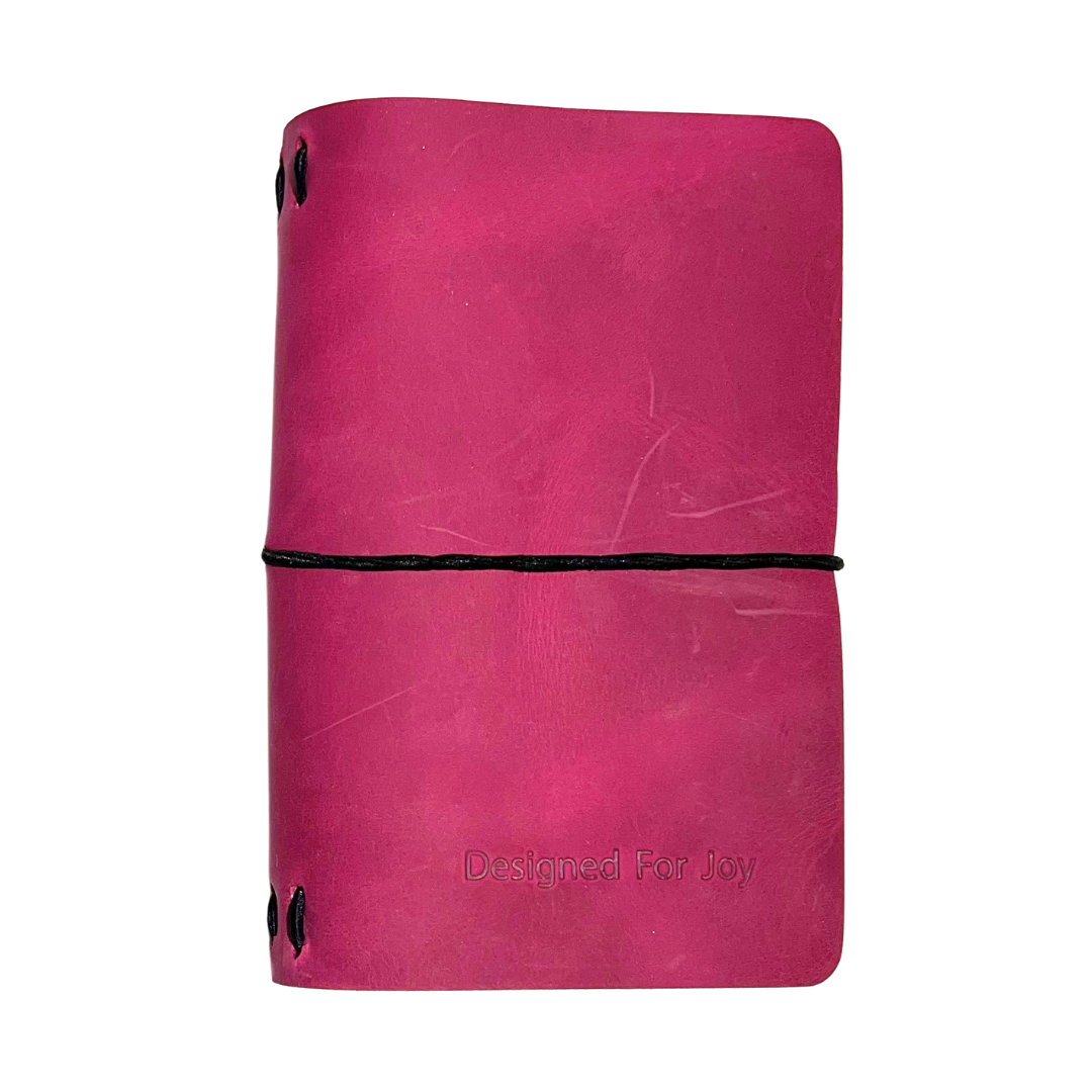 Pink notebook with 'Designed for Joy' text on the cover and a black strap, on a white background