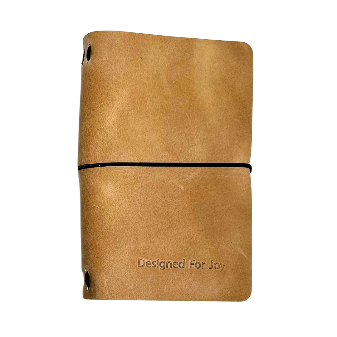 Light brown notebook with 'Designed for Joy' text on the cover and a black strap, on a white background