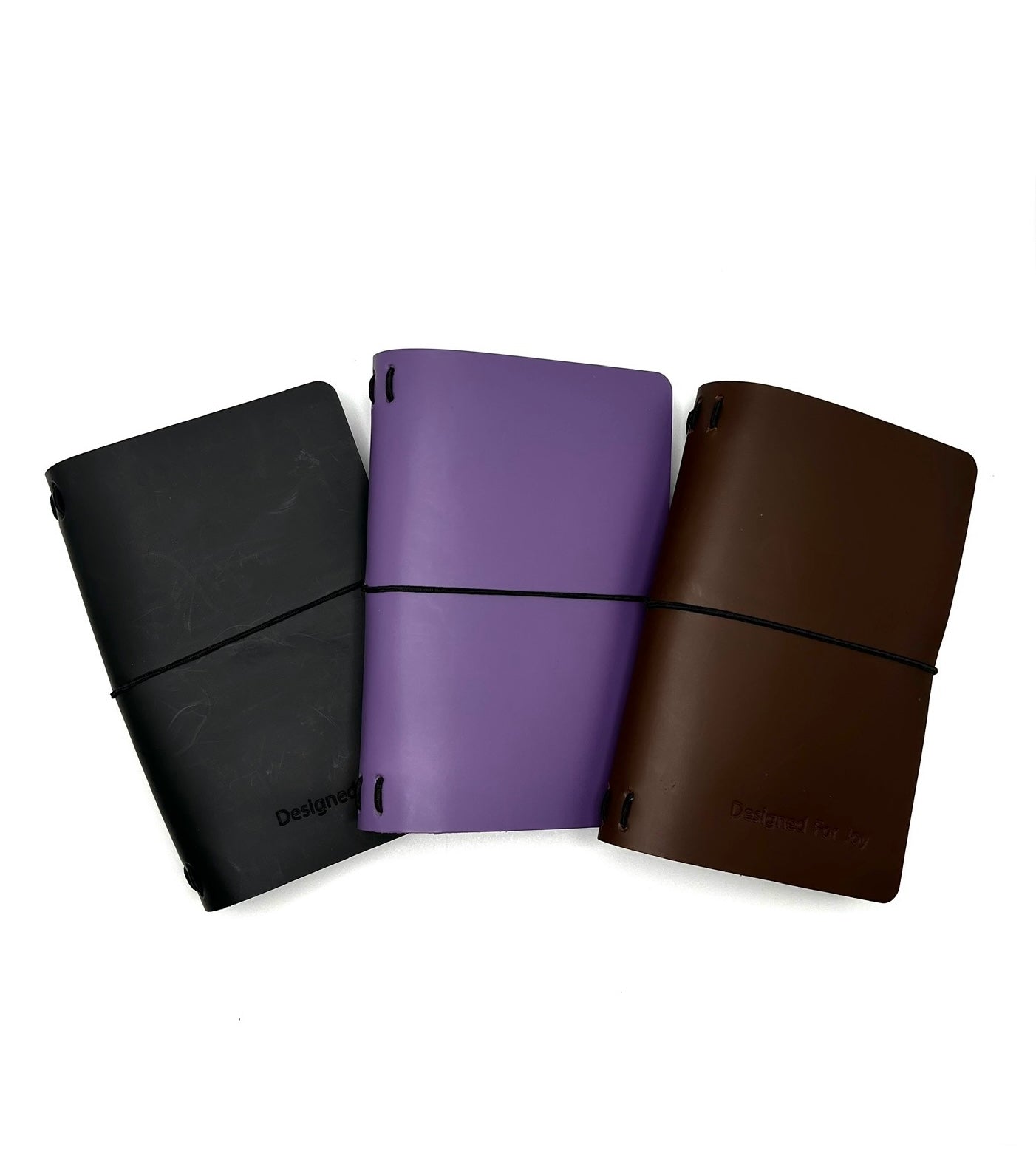 Three leather journals in black, purple, and dark brown colors, with 'Designed for Joy' text and a black strap on a white background
