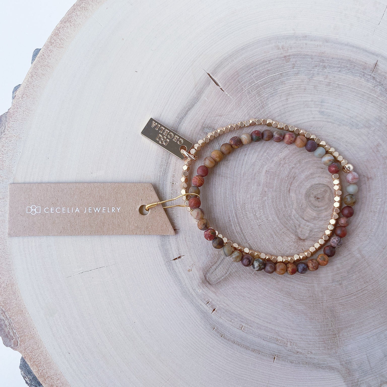A half red creek jasper, half matte gold-colored beaded bracelet on a wooden surface with a tag that has 'Cecelia Jewelry' text