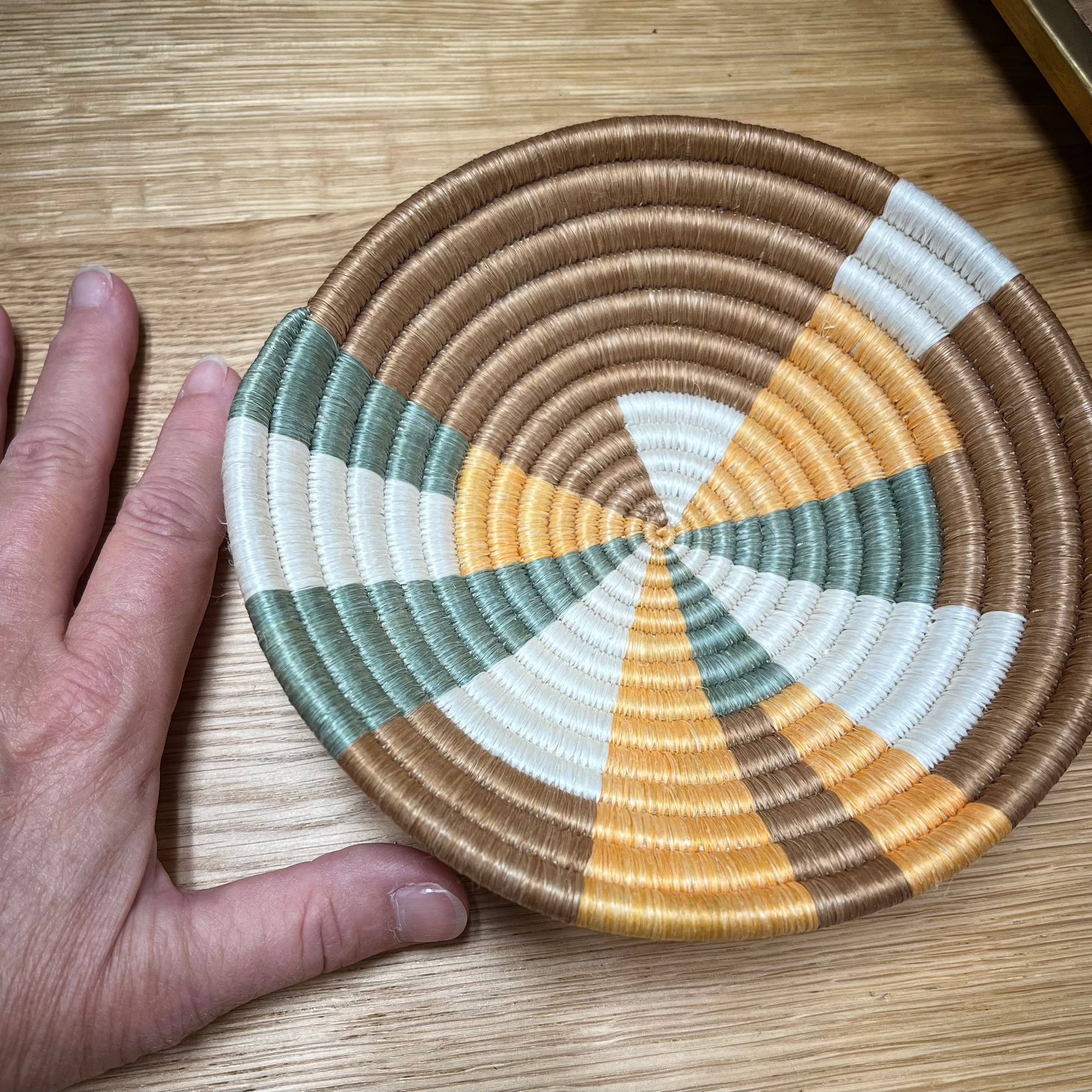 A small brown, orange, white, and green Rwandan basket, displayed on a wooden surface next to a hand