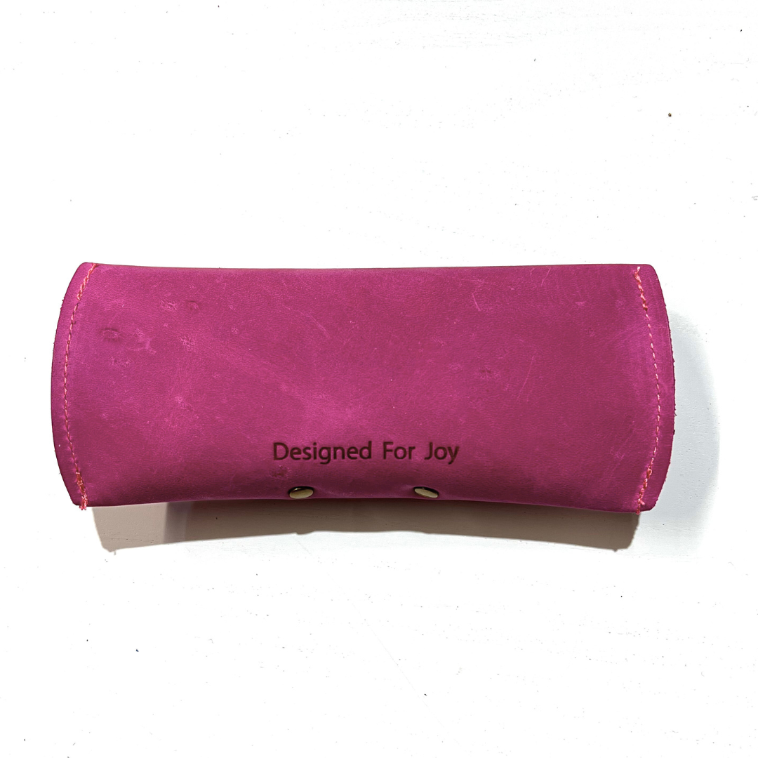 Back of a raspberry pink leather sunglasses case with 'Designed for Joy' text, displayed on a white background