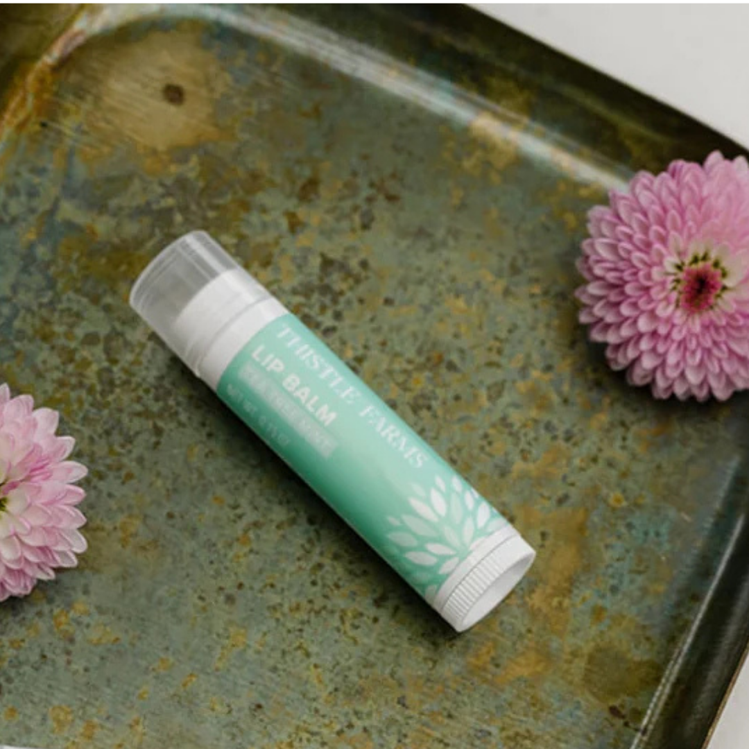A stick of 'Thistle Farms Lip Balm Tea Tree Mint' on a rusty tray with two pink flowers