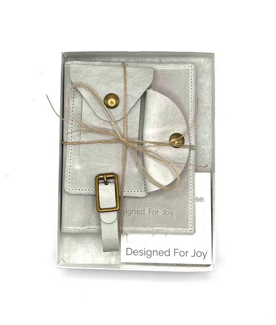 Gray leather travel bundle including a passport cover, a cord organizer, and a luggage tag, packaged for gifting with a card that reads 'Designed For Joy'