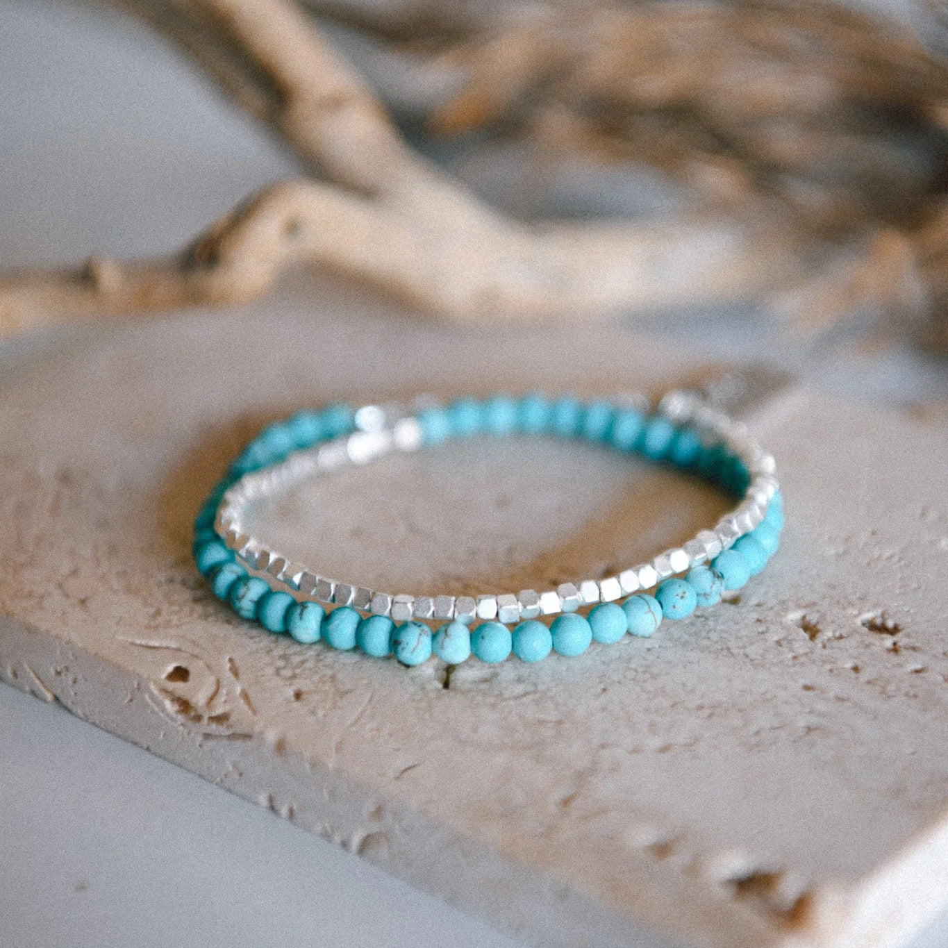 A half turquoise, half matte silver-colored beaded bracelet on a stone surface with a blurred background